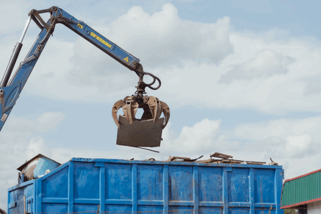 A crane scrap grabber loads the old metal into an industrial dumpster at the recycling plant.