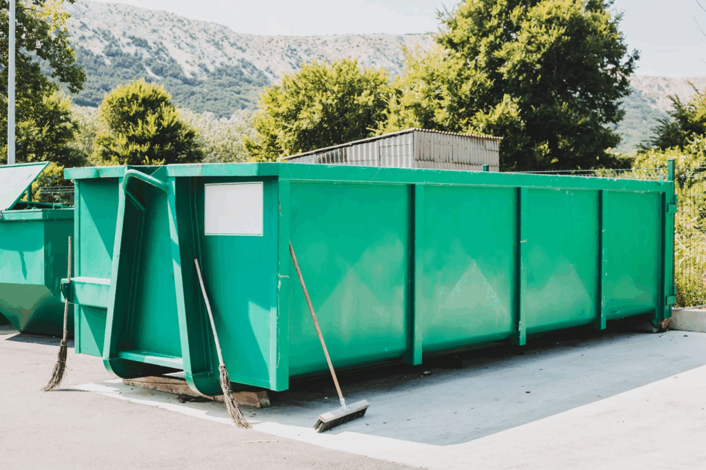 Commercial dumpsters next to brooms and trees.