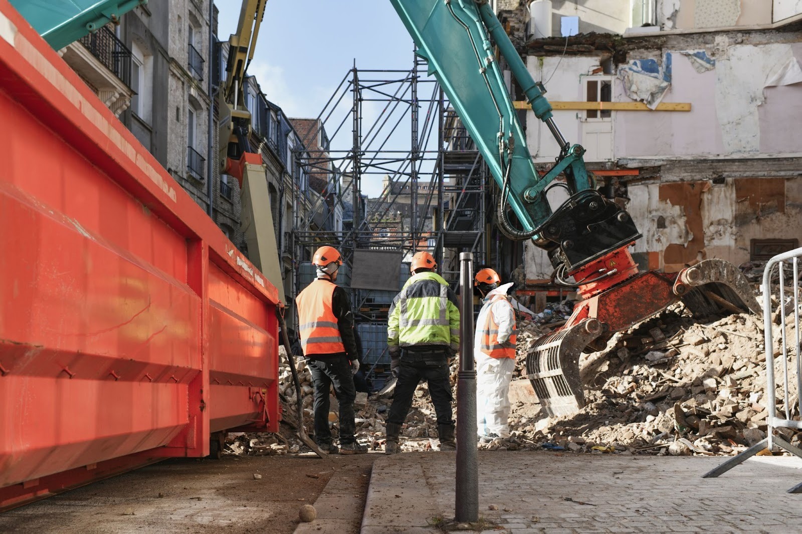 Large orange demolition dumpster beside an active demolition site with workers observing